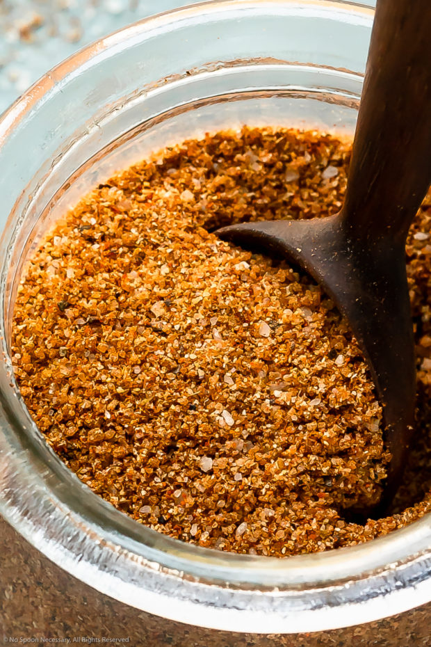 Angled, extreme close-up photo of BBQ Dry Rub in a glass jar with a spoon inserted into the rub.