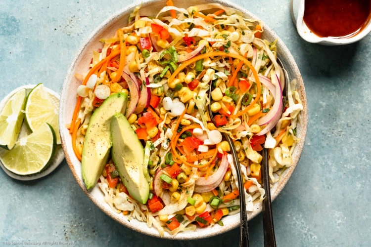 Overhead photo of Mexican coleslaw with corn in a white bowl with two serving spoons inserted into the slaw and a ramekin of fresh lime wedges and jar of vinaigrette dressing next to the bowl.