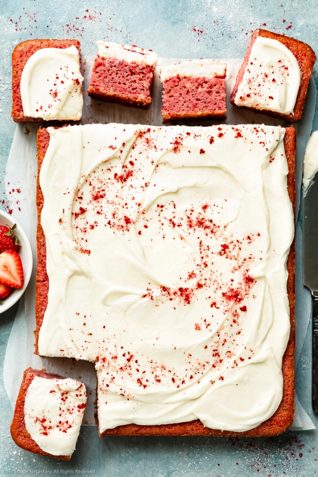 Overhead photo of Strawberry Sheet Cake topped with cream cheese frosting and dusted with crushed dried strawberries on a blue surface; with a few squares of cake cut and pulled away from the cake.