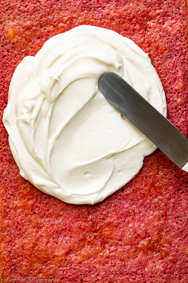 Overhead, up close photo of a strawberry cake being slathered with cream cheese frosting with an offset spatula - photo of step 13 of the recipe.