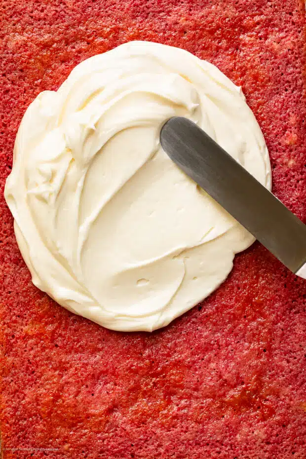 Action photo of an offset spatula spreading cream cheese frosting on a pink cake.