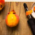 Overhead photo of a partially peeled peaches on a cutting board with a vegetable peeler next the to fruit.