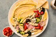 Overhead photo of a hummus bowl with greek chicken, cucumbers, pickled onions, cherry tomatoes, and kalamata olives.