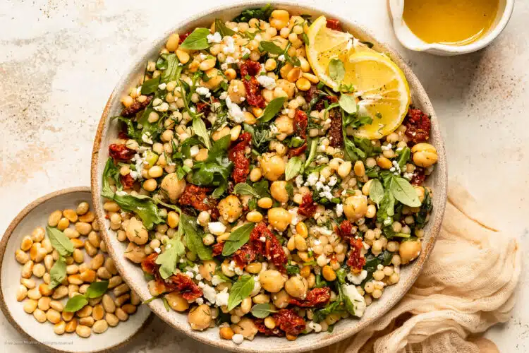 Overhead landscape photo of pearl couscous with sun-dried tomatoes, arugula and chickpeas in a large white bowl with a jar of lemon vinaigrette next to the bowl.