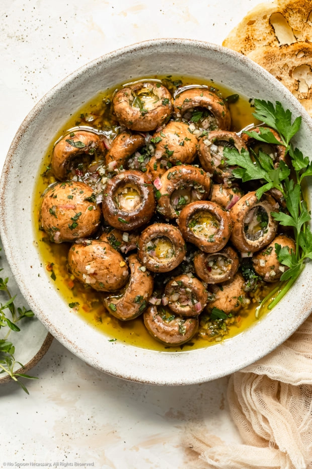 Overhead photo of homemade marinated mushrooms garnished with fresh parsley in a large white serving bowl with slices of artisan bread and a ramekin of fresh thyme next to the bowl.