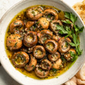 Overhead landscape photo of homemade marinated mushrooms garnished with fresh parsley in a large white serving bowl with slices of artisan bread and a ramekin of fresh thyme next to the bowl.