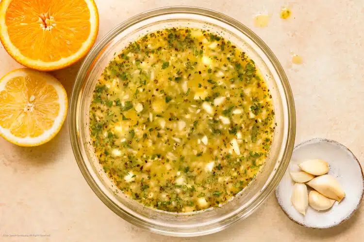 Photo of mojo marinade in a glass mixing bowl with an orange, lemon, and garlic cloves next to the bowl.