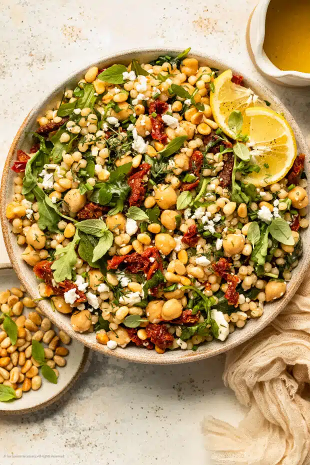 Overhead photo of Israeli couscous with sun-dried tomatoes, arugula and chickpeas in a large white bowl with a jar of lemon vinaigrette next to the bowl.