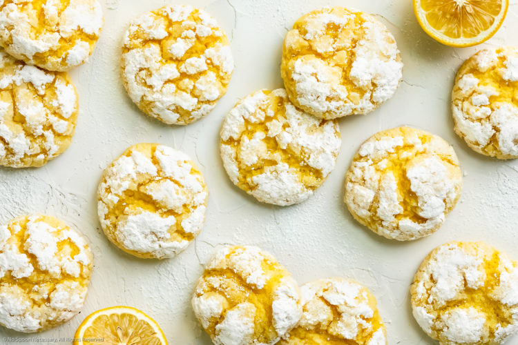 Overhead landscape photo of a dozen Cake Mix Lemon Crinkle Cookies topped with powdered sugar haphazardly arranged on a white wooden board, with fresh halves of lemon scattered around the cookies.