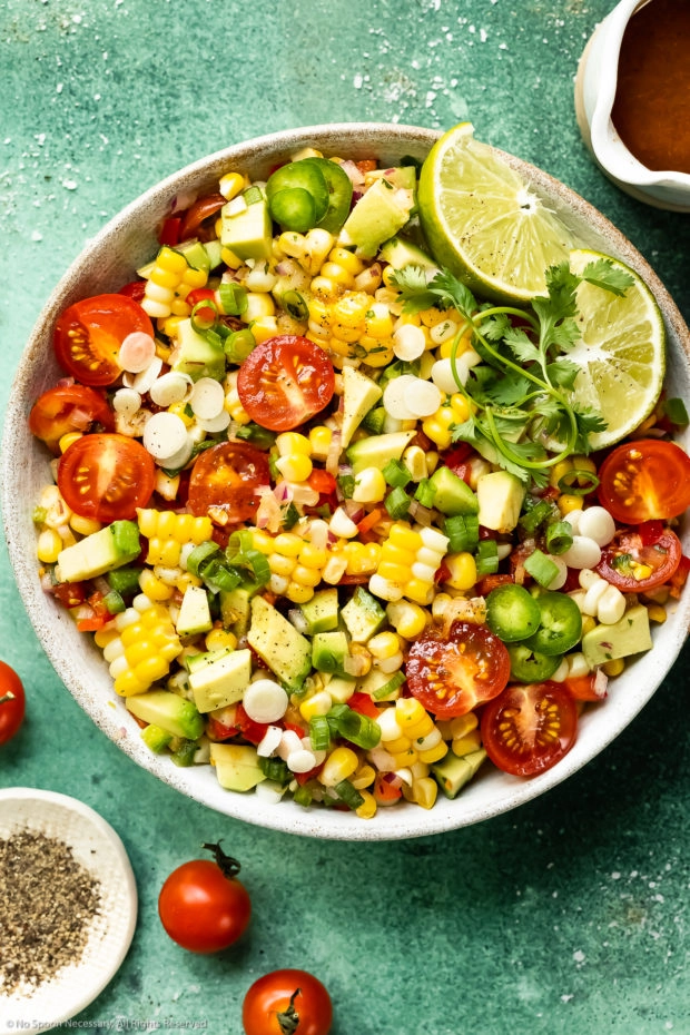 Overhead photo of Mexican Corn Salad with tomatoes and avocados in a white serving bowl with a jar of honey lime vinaigrette and a ramekin of fresh ground pepper arranged around the bowl.