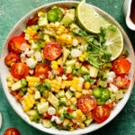 Overhead, landscape photo of Mexican Corn Salad with tomatoes and avocados in a white serving bowl with a jar of honey lime vinaigrette and a ramekin of fresh ground pepper arranged around the bowl.