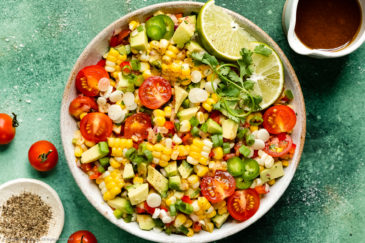 Overhead, landscape photo of Mexican Corn Salad with tomatoes and avocados in a white serving bowl with a jar of honey lime vinaigrette and a ramekin of fresh ground pepper arranged around the bowl.