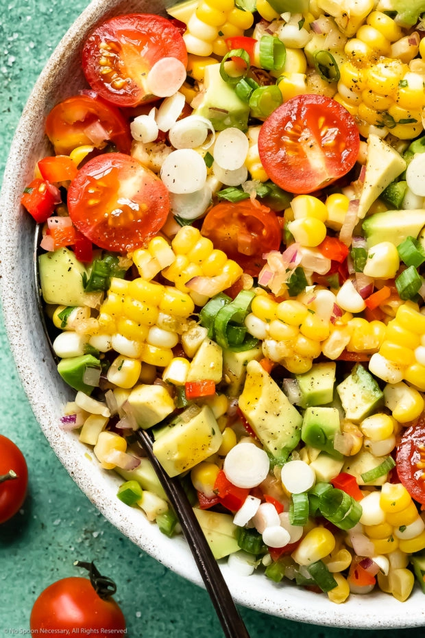 Overhead close-up photo of Corn Tomato Avocado Salad in a white bowl with a serving spoon tucked into the salad.