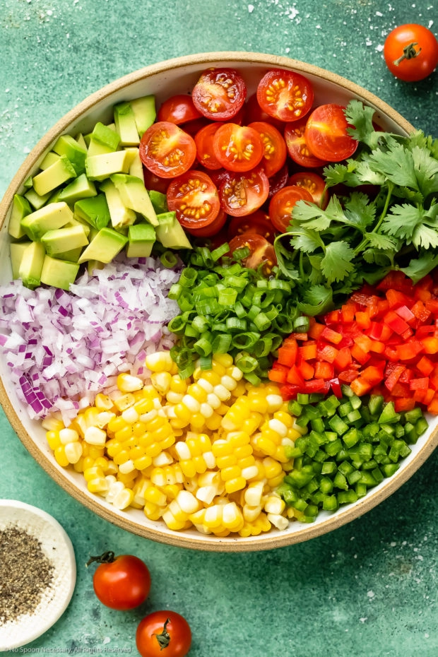 Overhead photo of all the ingredients needed to make sweet summer corn salad neatly arranged in a large bowl with a ramekin of freshly ground pepper next to the bowl.