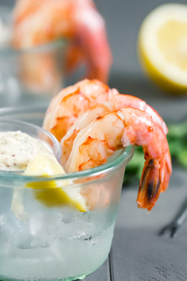 Straight on photo of a small glass bowl filled with ice with three cocktail shrimp looped around the edges of the bowl and a small ramekin of Remoulade on top of the ice.