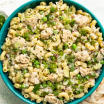 Overhead photo of prepared Tuna Macaroni Pasta Salad with peas and fresh herbs in a teal serving bowl with a ramekin of sliced scallions and pale tan napkin next to the bowl.