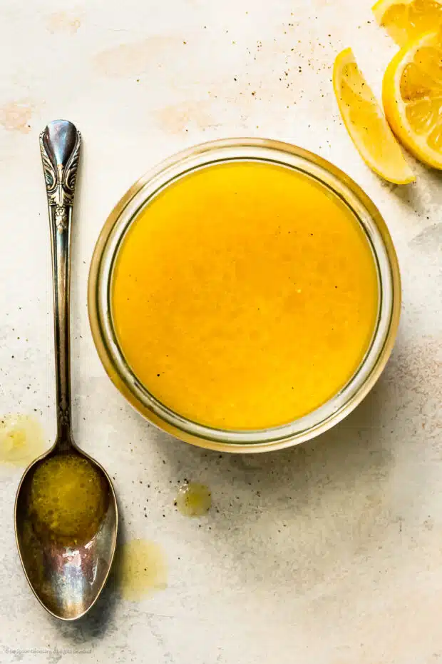 Overhead photo of lemon vinaigrette in a glass jar with a mixing spoon and fresh lemon wedges next to the jar.