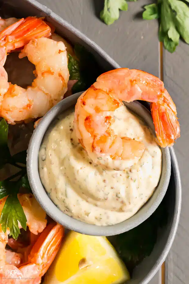 Overhead close-up photo of a poached shrimp cocktail dipped into a ramekin of remoulade sauce.