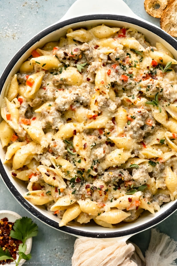 Overhead close-up photo of creamy sausage pasta garnished with fresh parsley in a large white skillet with ramekin of crushed red pepper flakes next to the skillet.