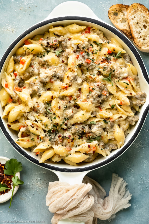 Overhead photo of creamy Italian sausage pasta garnished with fresh parsley in a large white skillet with slices of crusty bread and a ramekin of crushed red pepper flakes next to the skillet.