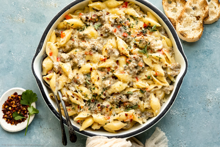 Overhead, landscape photo of creamy Italian sausage pasta garnished with fresh parsley in a large white skillet with slices of crusty bread and a ramekin of crushed red pepper flakes next to the skillet.
