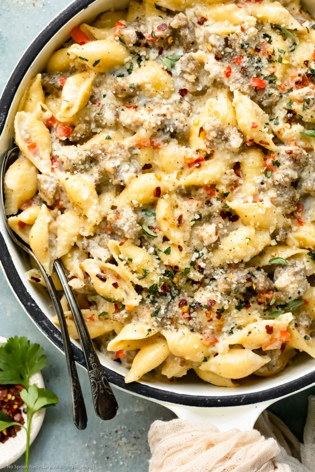 Overhead photo of creamy pasta with spicy Italian sausage in a large white skillet with two serving spoons inserted into the pasta and a ramekin of crushed red pepper flakes next to the skillet.
