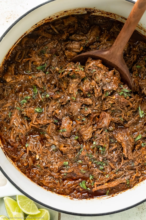 Overhead photo of juicy Mexican shredded beef in a large white pot with a wooden serving spoon inserted into the beef and a ramekin of fresh lime wedges next to the pot.