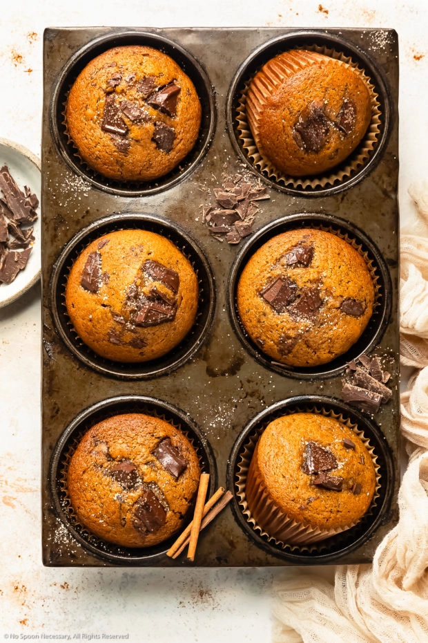 Overhead photo of pumpkin chocolate chip muffins in a muffin pan with roughly chopped dark chocolate and cinnamon sugar strewn on top of the pan and a pale tan napkin next to the muffins.