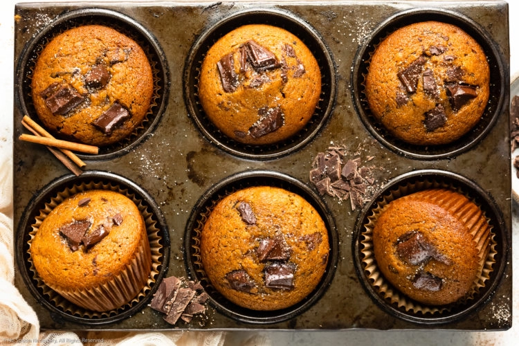 Overhead landscape photo of pumpkin chocolate chip muffins in a muffin pan with roughly chopped dark chocolate and cinnamon sugar strewn on top of the pan.
