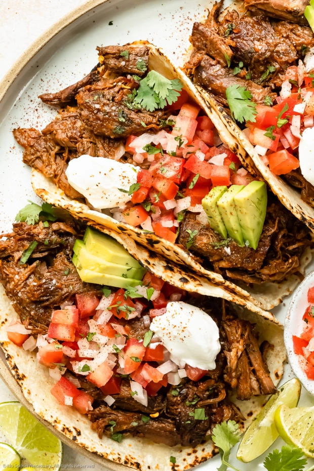 Overhead photo of Mexican Beef Tacos topped with fresh pico de gallo, sliced avocados and sour cream on a white plate with lime wedges next to the tacos.