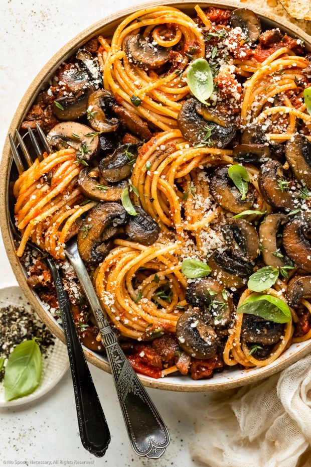 Overhead, close-up photo of a bowl of Mushroom Spaghetti garnished with fresh basil leaves and grated parmesan with a serving fork and spoon inserted into the pasta and a ramekin of crushed black pepper next to the bowl.