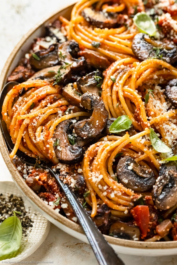 Angled, close-up photo of a bowl of Mushroom Spaghetti with a serving spoon inserted into the pasta and a ramekin of crushed black pepper next to the bowl.
