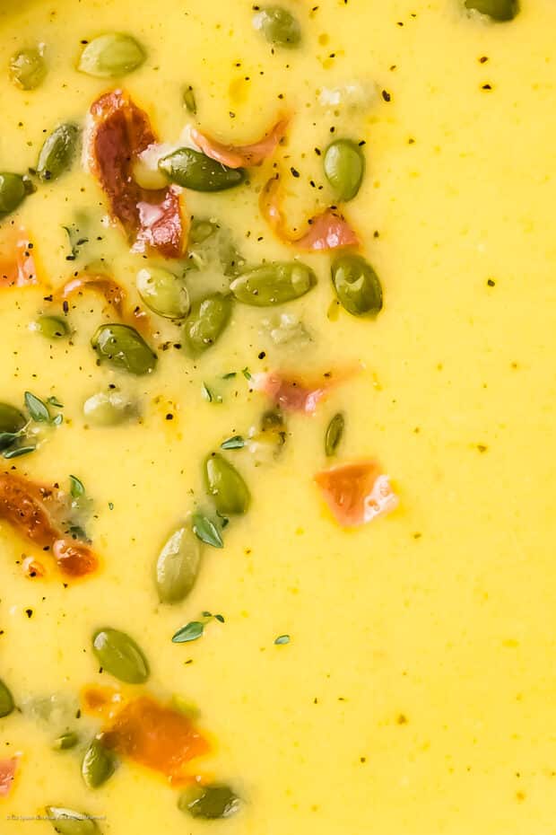 Close-up photo of a yellow colored soup with garnished with crispy bacon and toasted pumpkin seeds.
