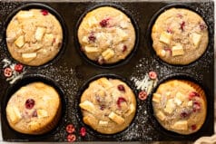 Overhead landscape photo of Chocolate Cranberry Muffins dusted with powdered sugar in a muffin pan with a ramekin of fresh cranberries, glass of milk and pale tan napkin next to the pan.