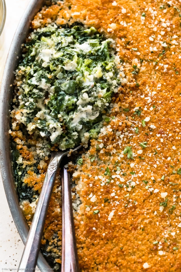 Slightly angled, close-up photo of Creamed Spinach Casserole in an oval baking pan with two serving spoons tucked into the casserole exposing the creamy spinach interior.