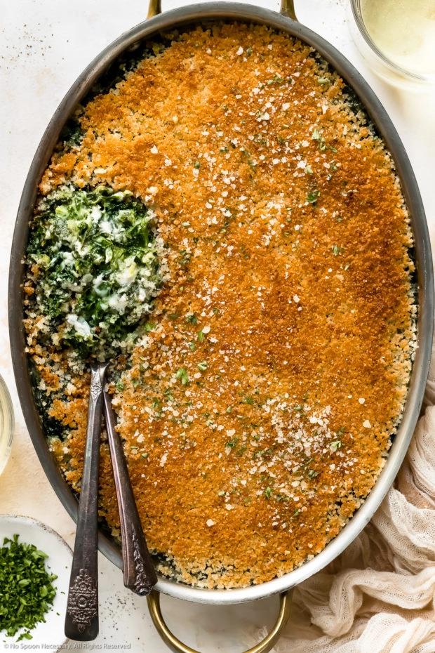 Overhead photo of Creamed Spinach Casserole in an oval baking pan with two serving spoons tucked into the casserole exposing the creamy spinach interior.