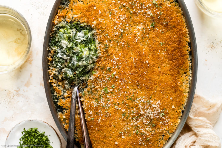 Overhead photo of Creamed Spinach Casserole in an oval baking pan with two serving spoons tucked into the casserole exposing the creamy spinach interior; with two wine glasses and a ramekin of chopped parsley next to the dish.