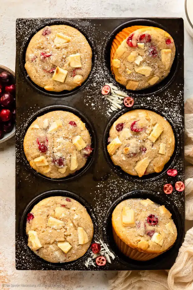 Overhead photo of Chocolate Cranberry Muffins dusted with powdered sugar in a muffin pan with a ramekin of fresh cranberries, glass of milk and pale tan napkin next to the pan.