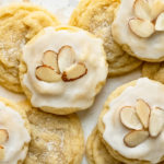 Overhead landscape photo of a spread out stack of soft and chewy almond sugar cookies on a white wood surface - the bottom cookies are plain while the cookies on top of the stack are covered with a cookie icing and decorative sliced almonds.