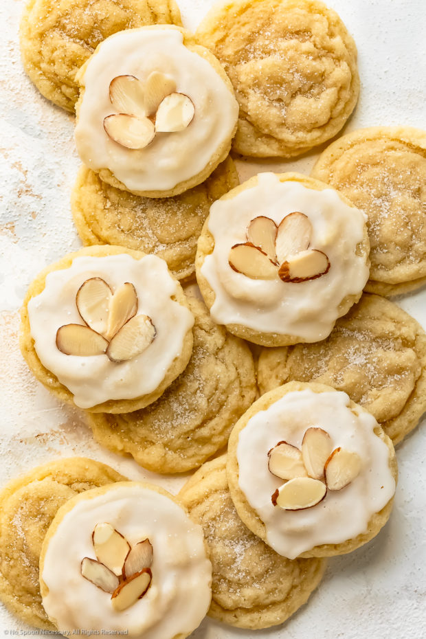 Overhead photo of a spread out stack of soft almond sugar cookies on a white wood surface - the bottom cookies are plain while the cookies on top of the stack are covered with a cookie icing and decorative sliced almonds.