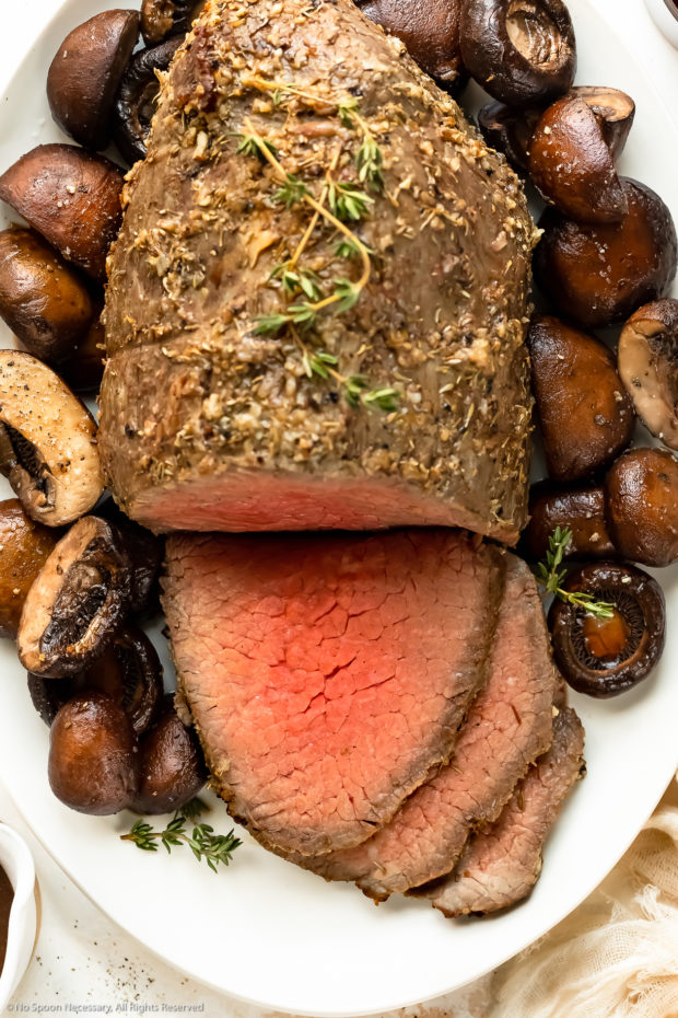 Overhead, close-up photo of a eye of round roast that's been partially sliced into to expose it's medium-rare center next to a bed of mushrooms on a white platter.