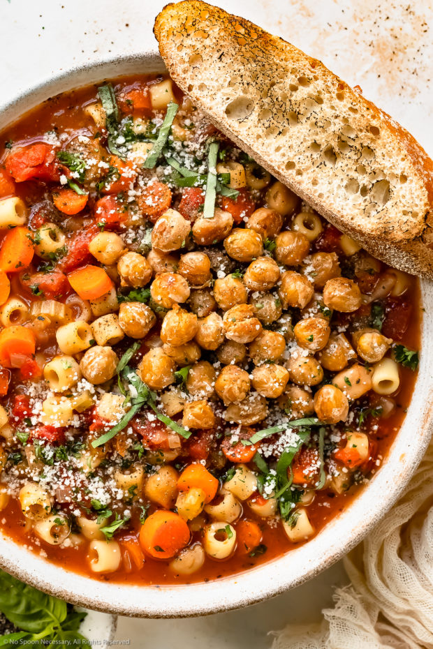 Overhead photo of Garbanzo Bean Soup garnished with crispy chickpeas, fresh basil and grated parmesan in a white bowl with a slice of artisan bread resting on the side of the bowl.