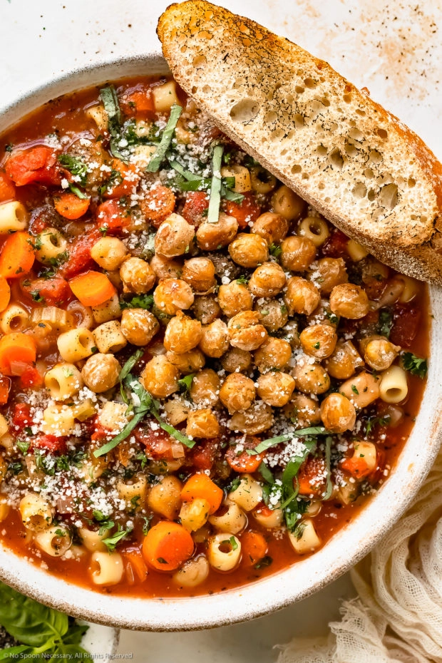 Overhead photo of Garbanzo Bean Soup topped with crispy chickpeas, fresh basil and grated parmesan in a white bowl with a slice of artisan bread resting on the side of the bowl.
