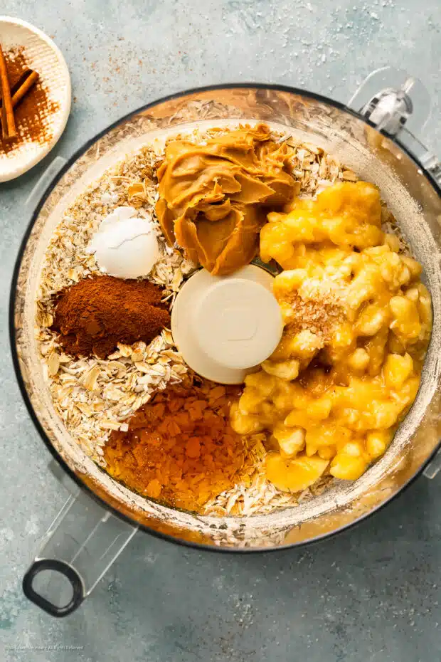 Overhead photo of a food processor bowl with rolled oats, mashed bananas, peanut butter, honey, and ground cinnamon.