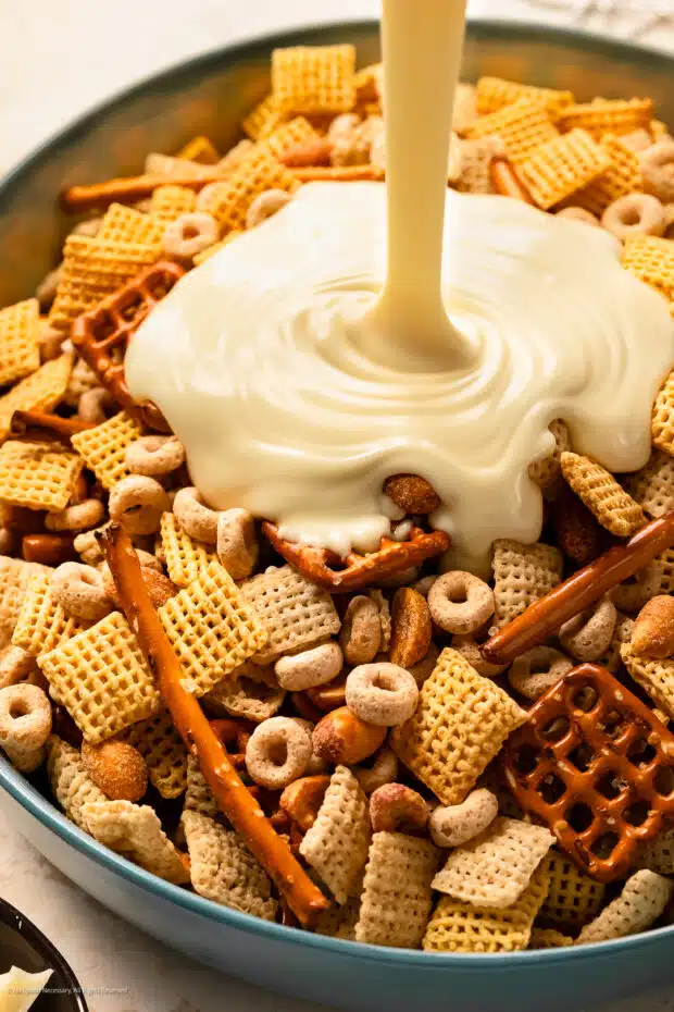 Action photo of melted white chocolate being poured over a sweet chex cereal mix with nuts and pretzels.