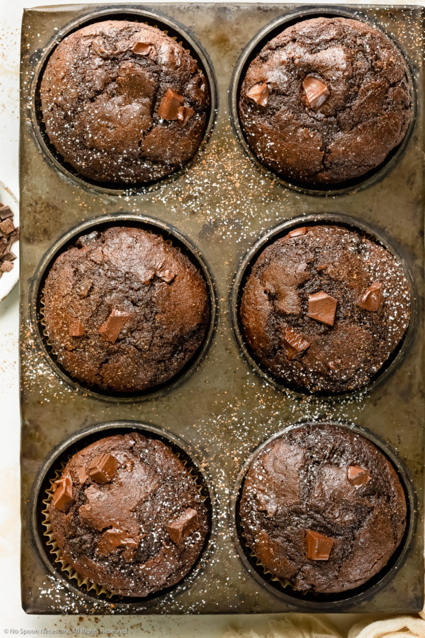 Overhead photo of fudgy chocolate muffins sprinkled with confectioner's sugar in a muffin baking pan.