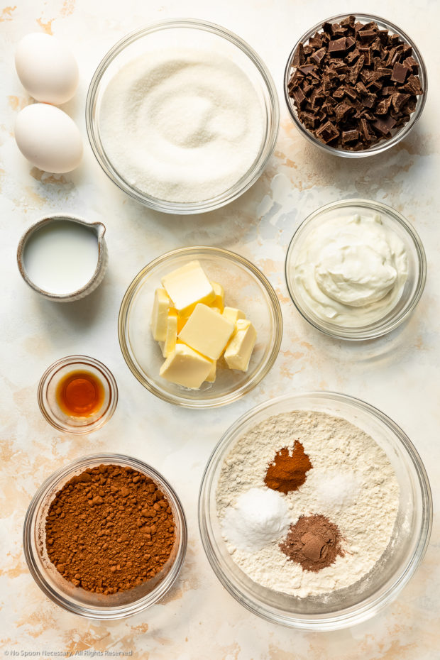 Overhead photo of all the ingredients needed to make chocolate chunk muffins neatly organized in individual bowls arranged on a white surface.