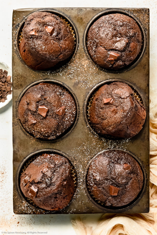 Overhead photo of double chocolate muffins in a muffin baking pan with a pale tan napkin and ramekin of chocolate chunks next to the baking pan.