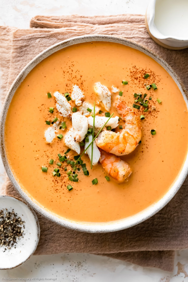 Overhead photo of easy bisque garnished with crab and shrimp in a white soup bowl with a ramekin of ground black pepper and a small container of cream next to the bowl.