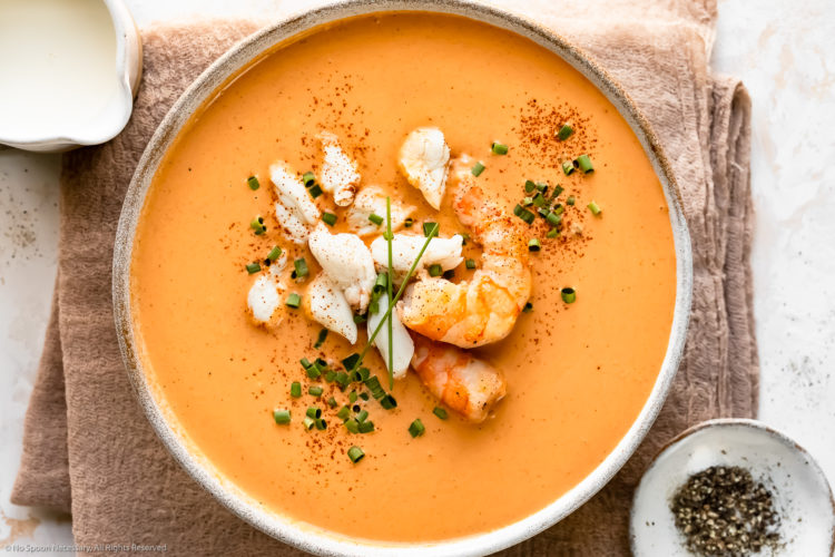 Overhead photo of Shrimp & Crab Seafood Bisque garnished with snipped chives in a white soup bowl with a ramekin of fresh cracked pepper and a small pitcher of cream next to the bowl.